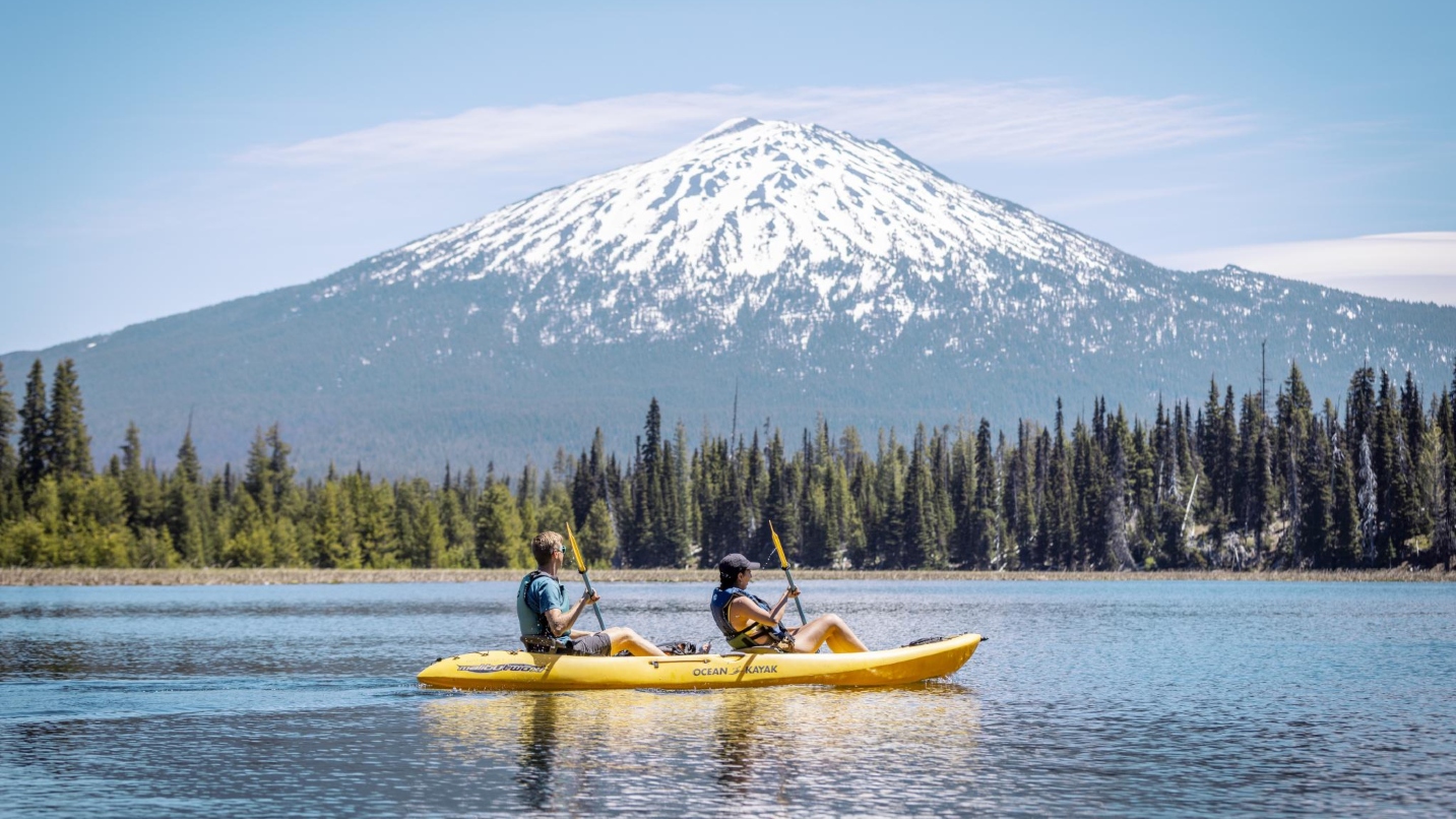 Man and woman sit in yellow kayak holding paddles on a calm lake with tall snow-capped mountain, trees and blue sky in background.