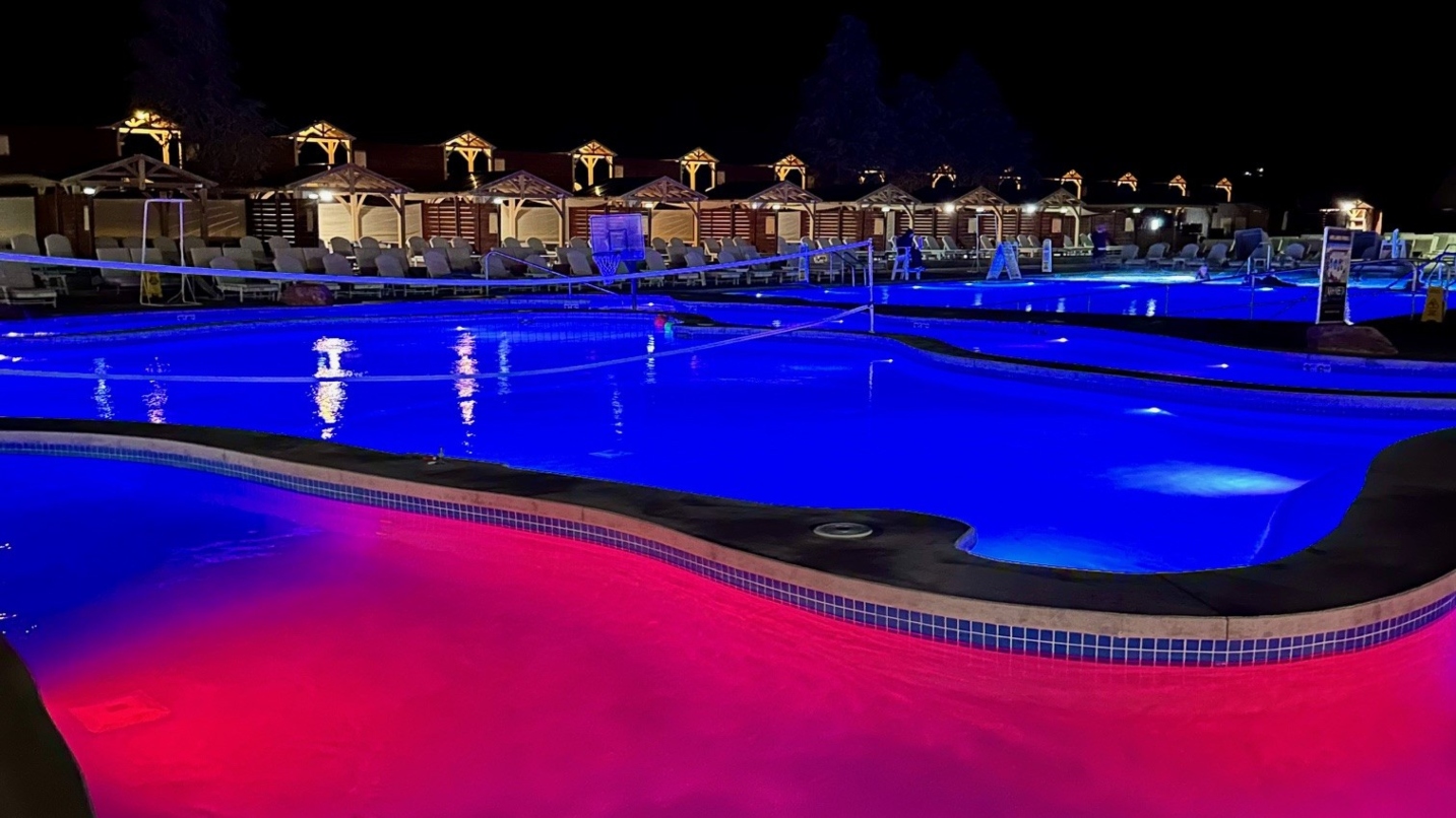 Lit-up blue and pink pools at a resort under a dark night sky, with cabanas and chairs to the left and a volleyball net over one of the pools.