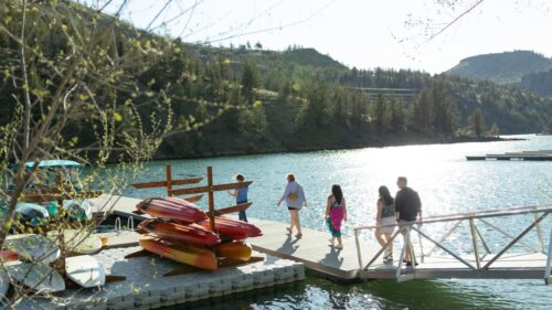 Five people walk down a walkway to a floating marina where a small boat is parked and several red kayaks and paddleboards are stacked. Trees and mountains are in the background and the lake is blue and sunny.