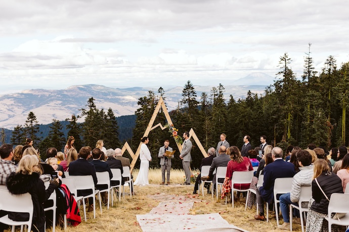 A bride and groom exchange vows with guests seated in folded chairs with a mountain landscape in the distance.