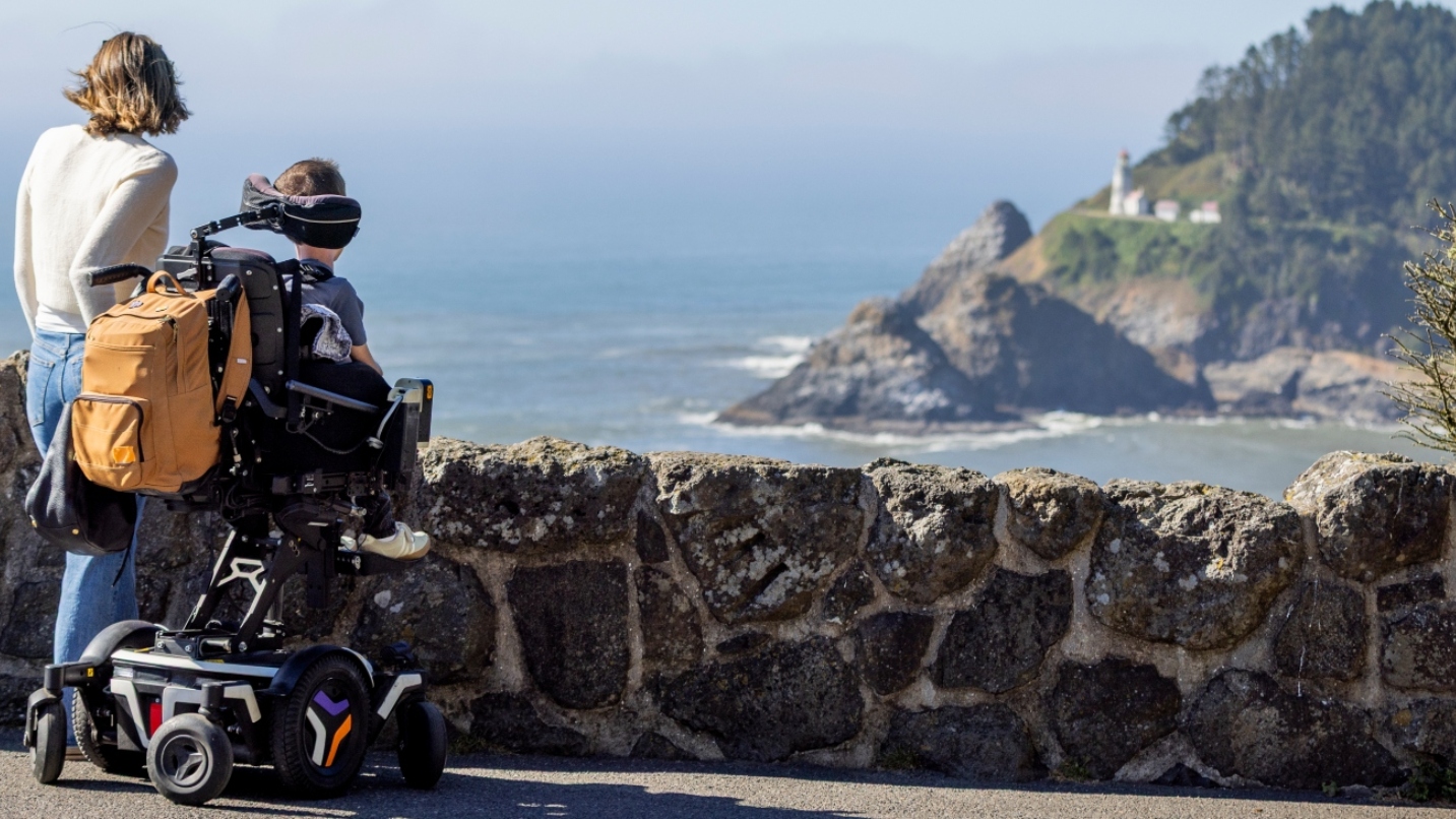 Woman stands next to man in wheelchair in front of a rock wall, both looking out to the sea and a lighthouse in the distance.