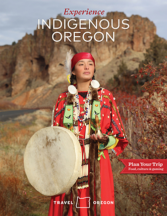 An indigenous woman wears a red coruroy trade dress and green beaded set, with wampum necklaces, bone and brass breastplate while she holds a hand drum and handbell.