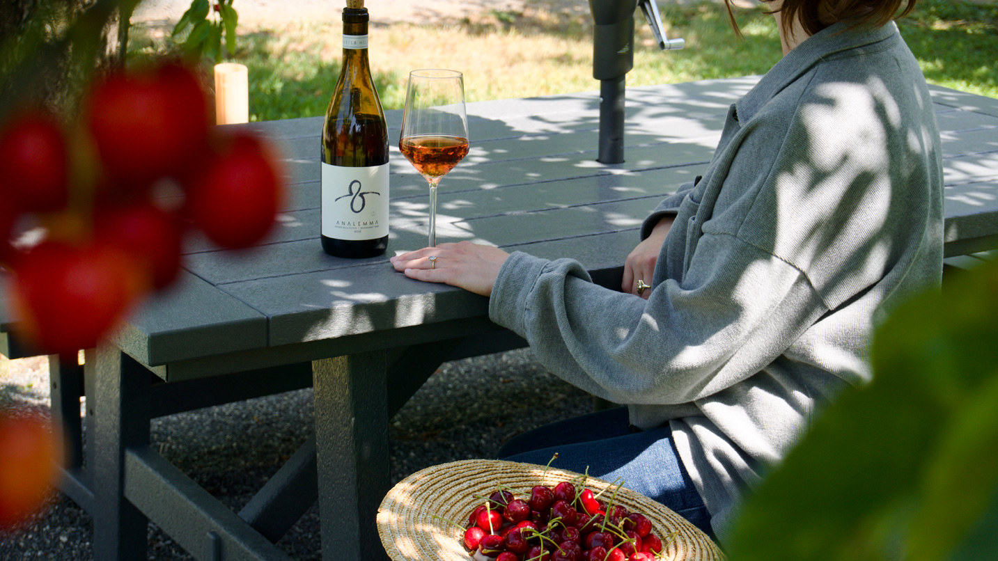 A person sitting at a picnic table with a glass of wine and bottle. Sitting next to her is a straw hat filled with cherries.