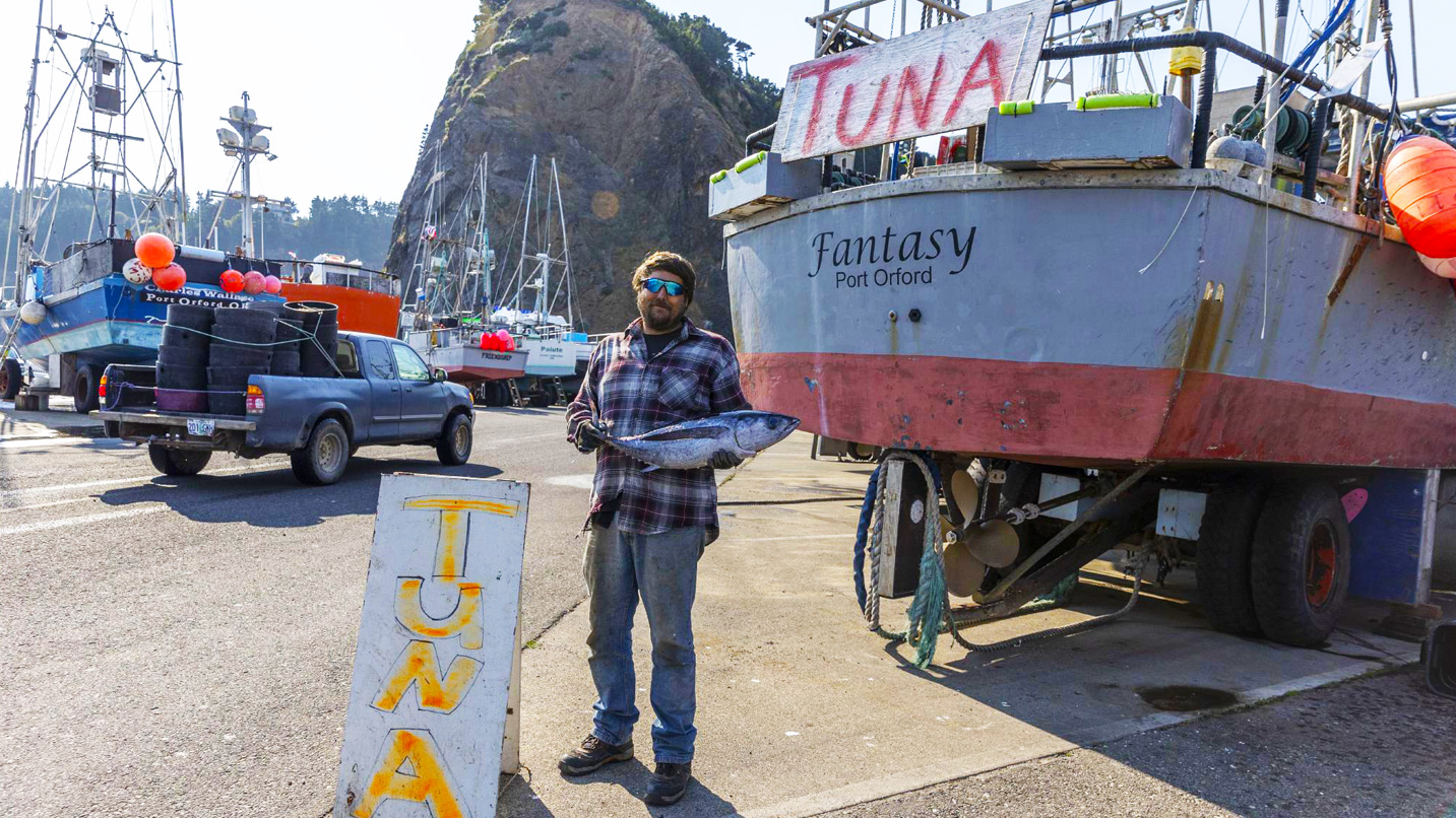 A man holding a small tuna fish beside a dory boat parked on land.