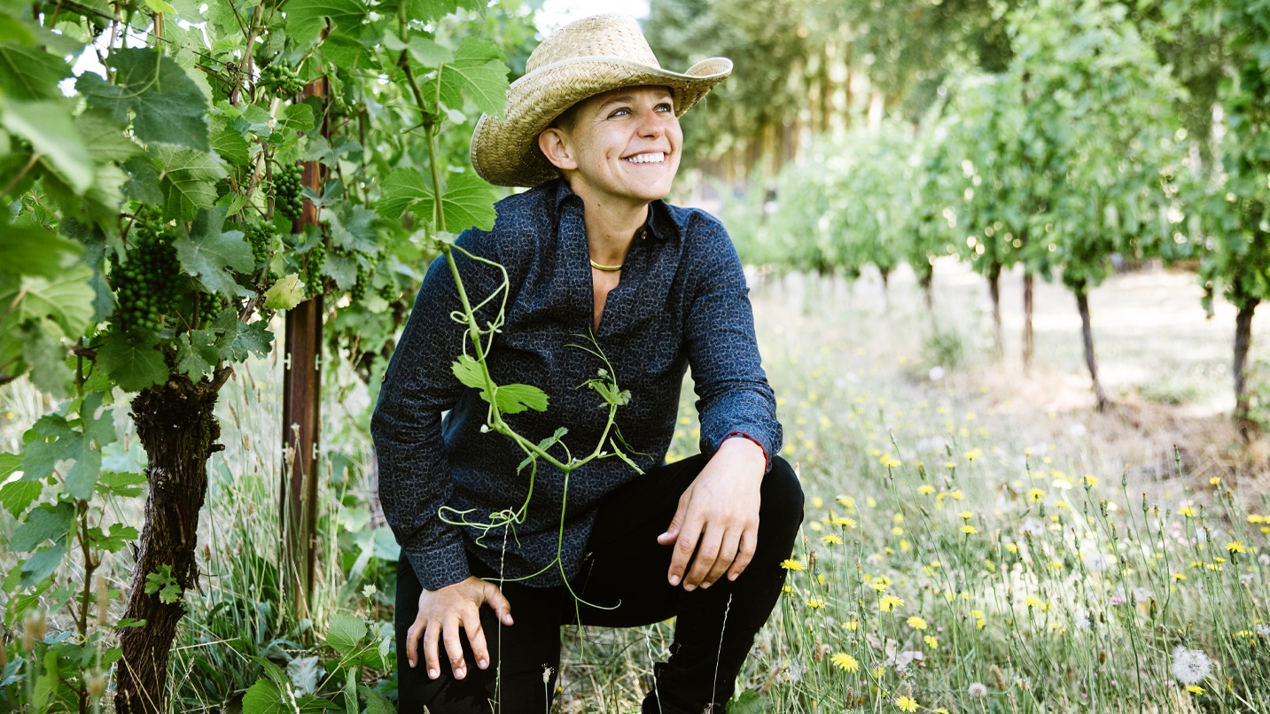 A woman kneeling next to grape vines.
