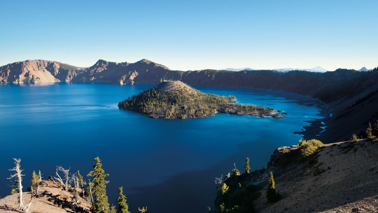 Large blue lake with a small island with trees in the middle, surrounded by caldera and trees, with blue sky above