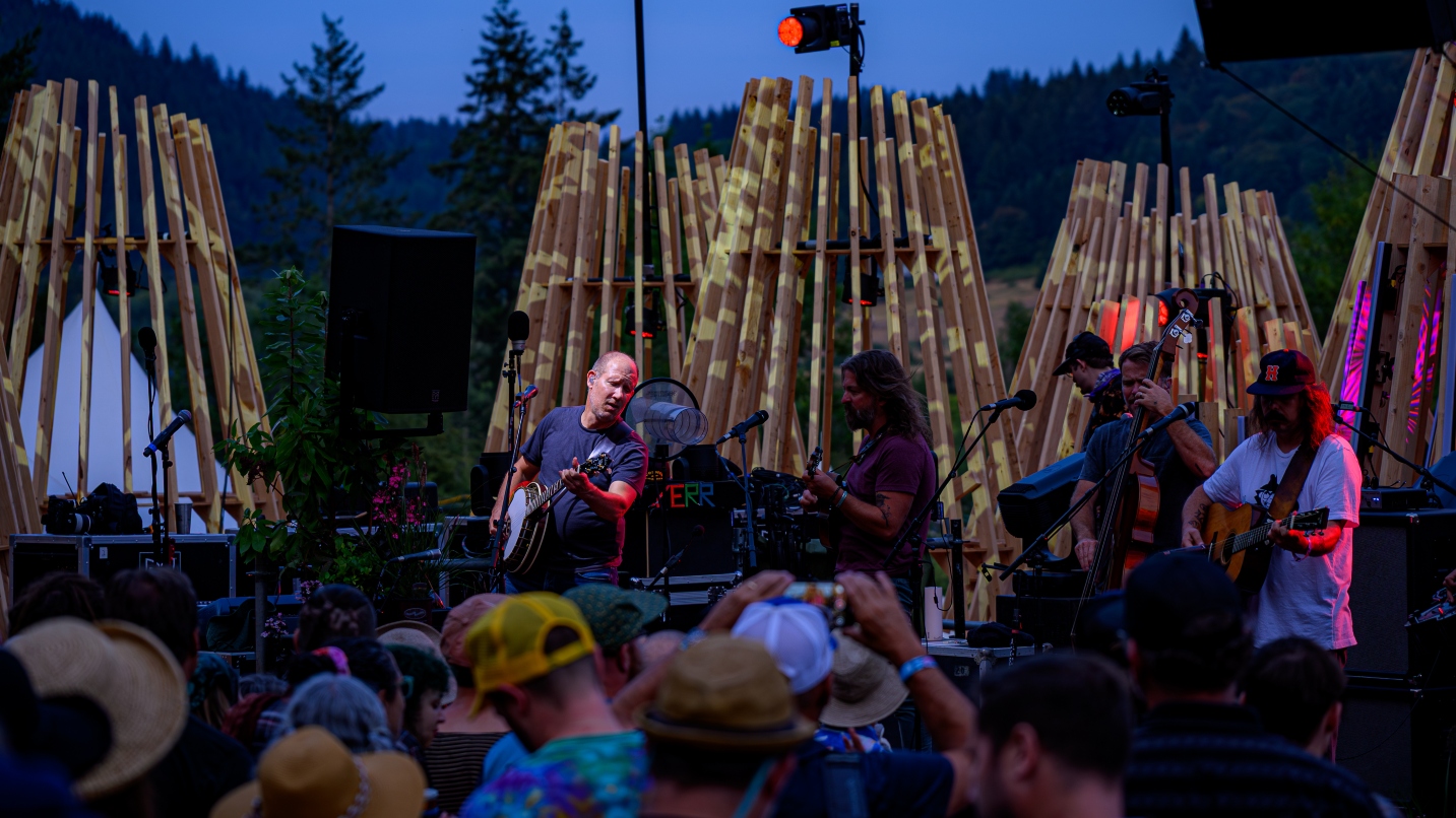 Musicians perform on stage against a night sky with wood-slat backdrops and people standing in the crowd
