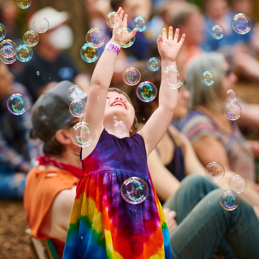 Small girl wearing tie-dye dress looks up at her arms at she tries to catch bubbles in a crowd of people