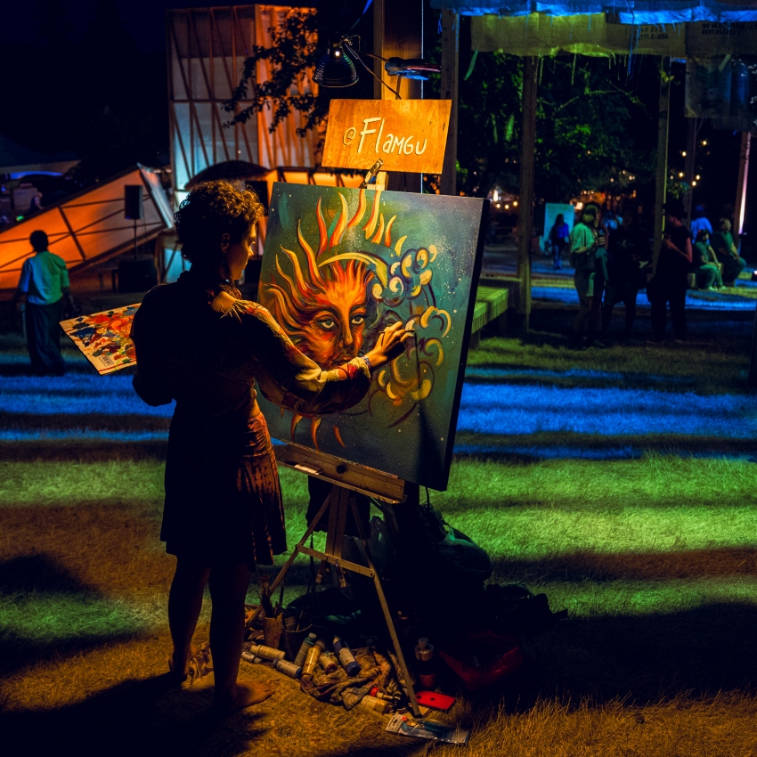 Woman with curly hair wearing dress paints a colorful painting with multi-colored lights shining on her under a night sky