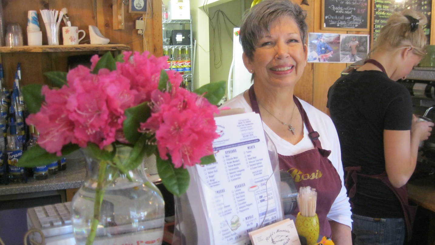 Woman with short grey hair and maroon apron smiles, next to a display of menus, vase of pink flowers and cash register.
