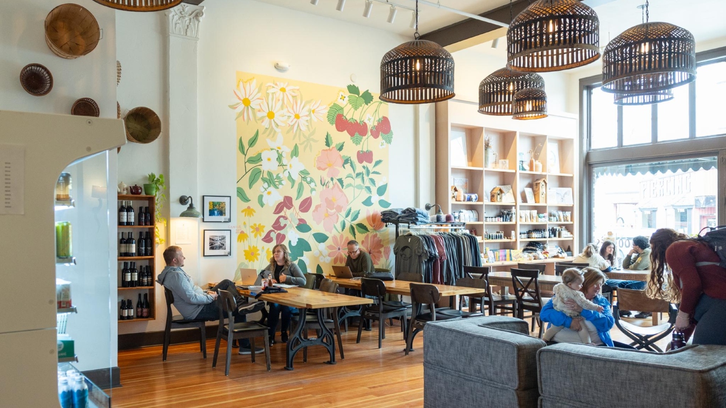 Hotel lobby with large windows, funky light fixtures, sofas, tables, chairs and people enjoying the space. Also a large yellow and red mural of fruit and plants on the wall.