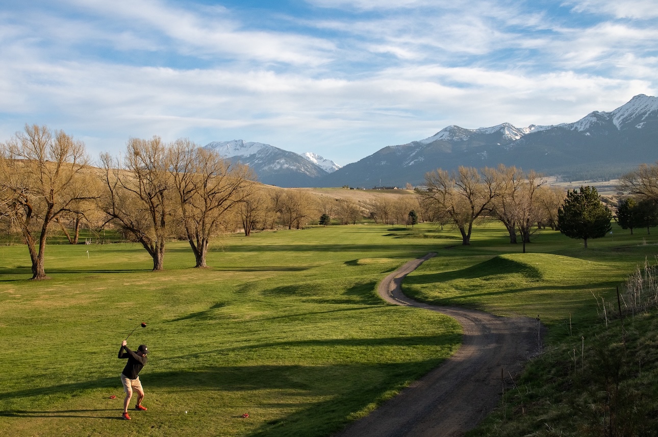 A fall scene as a golfer tees off with the greens extending to snow capped mountains in the distance.