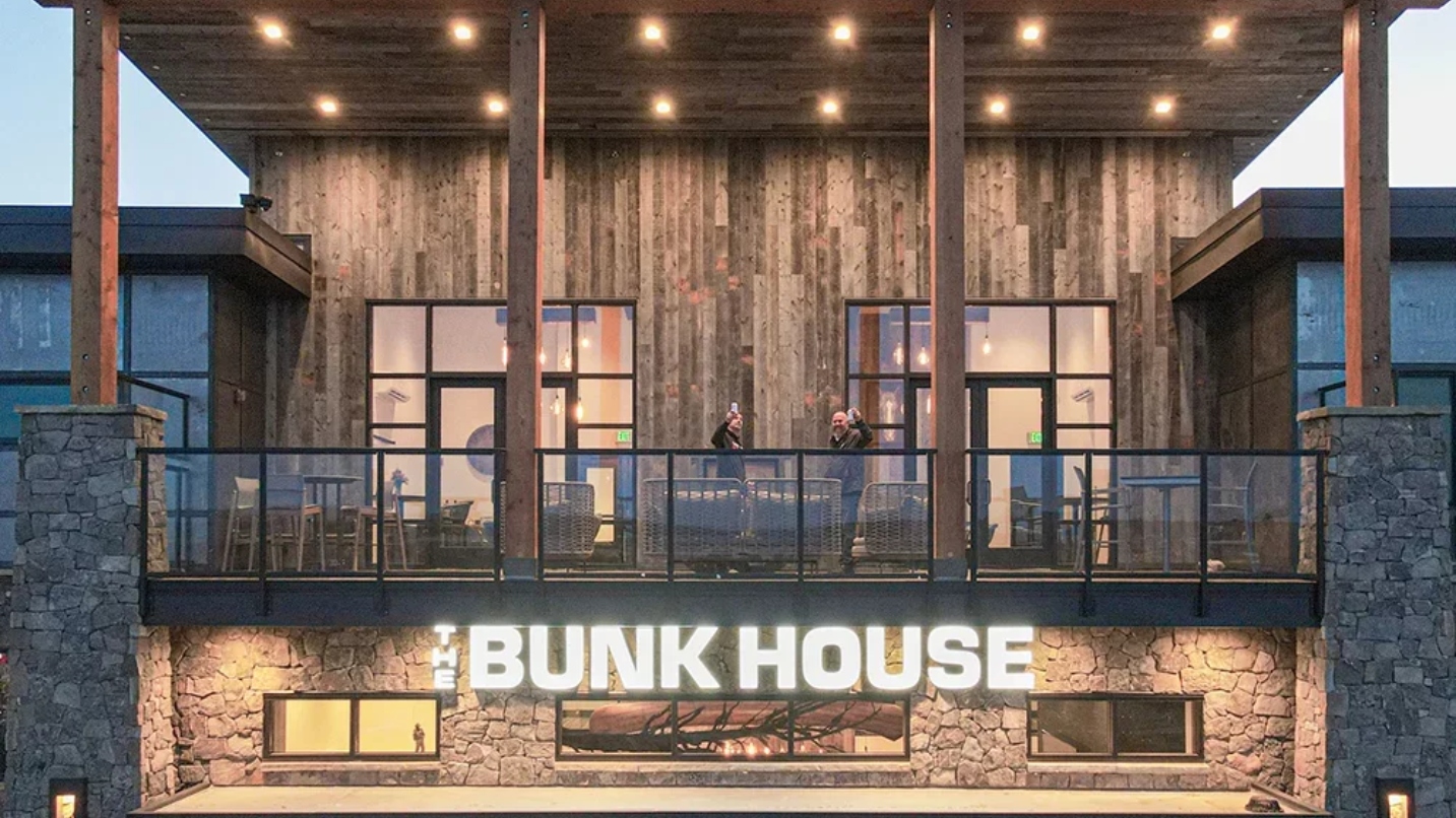 Two people stand on the lit balcony of a wood and stone building called the Bunk House and raise beverages in a toast
