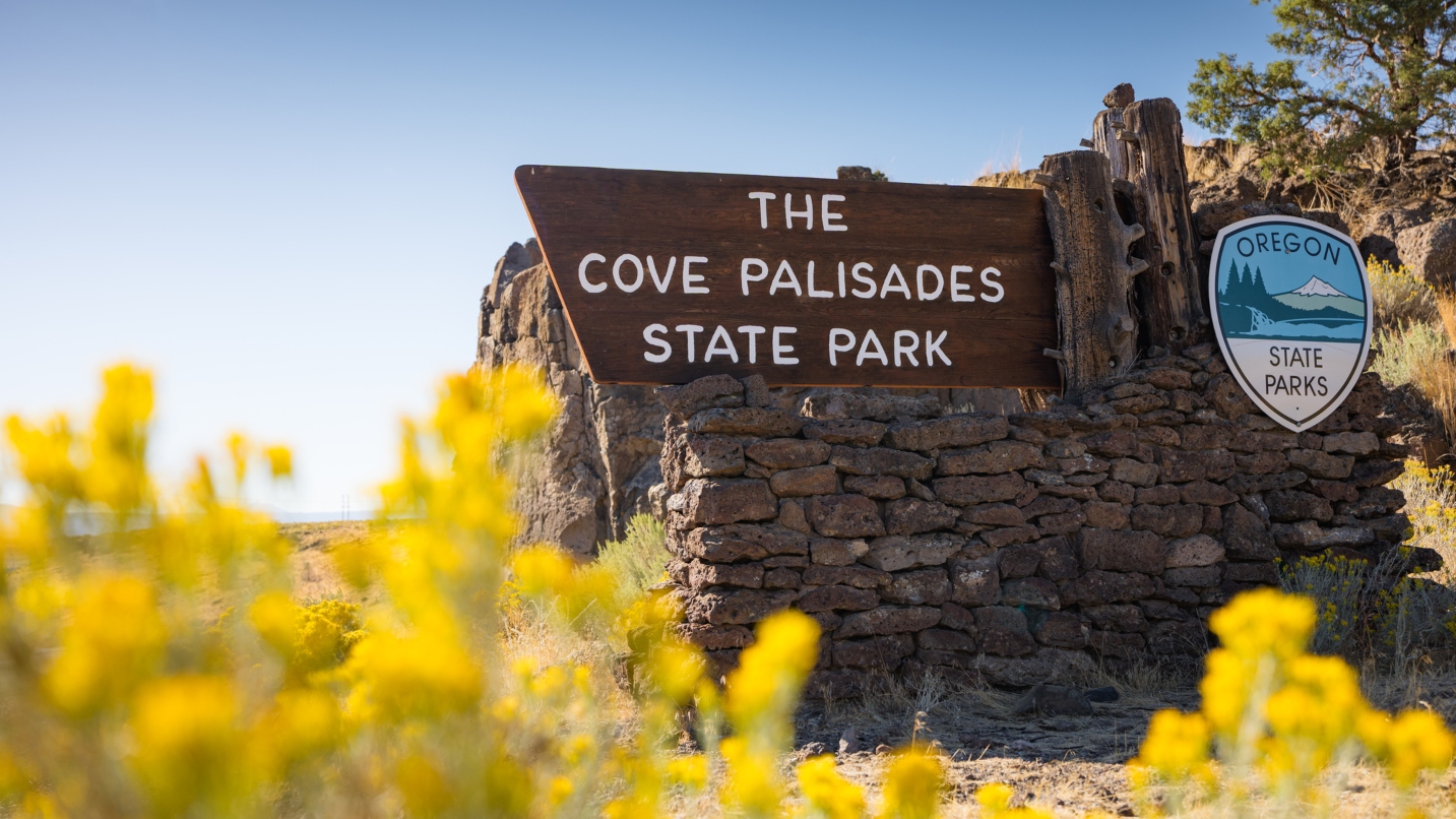 Brown brick and wood Oregon State Parks sign that reads "The Cove Palisades States Park" with blue sky and yellow shubs in foreground