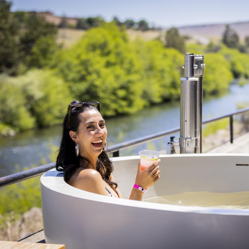 Woman with black hair and sunglasses on head smiles and holds a drink cup while sitting in hot tub, river landscape in background.