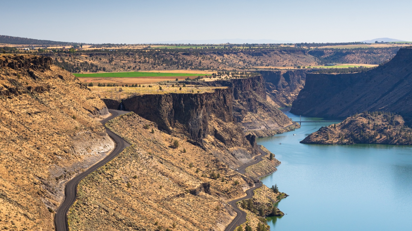 Steep canyon walls with plateaus and shrubs, with blue lake below, bridge in the distance and blue sky above