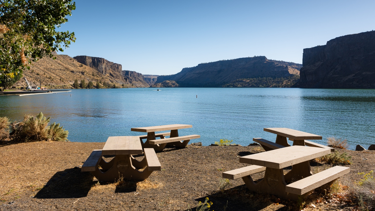 Large blue lake surrounded by canyon walls, with four picnic tables in a circle in the foreground on dirt ground.