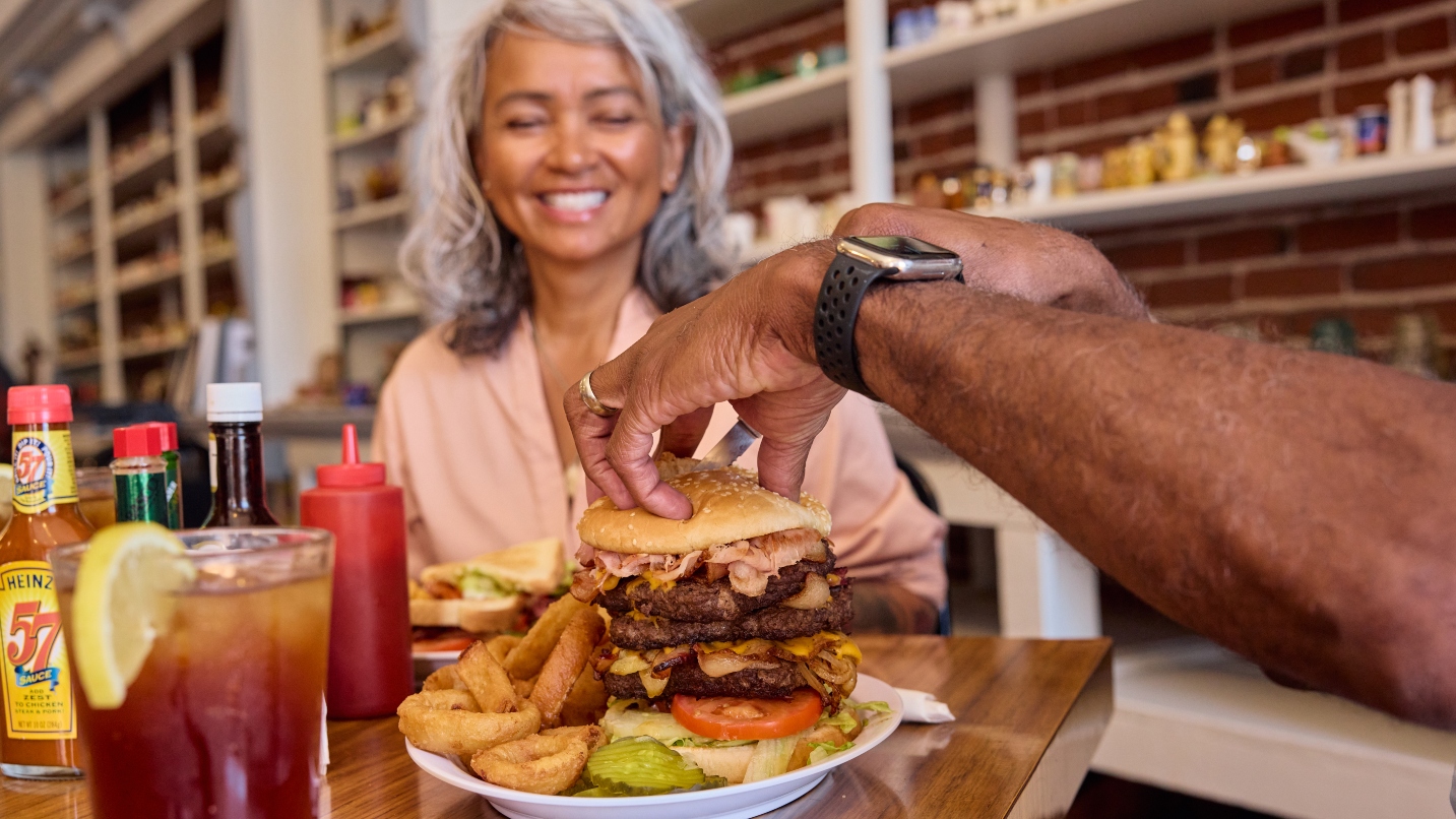 Woman with gray hair smiles at a close-up of a giant hamburger with all the fixings, held together by a man's arms