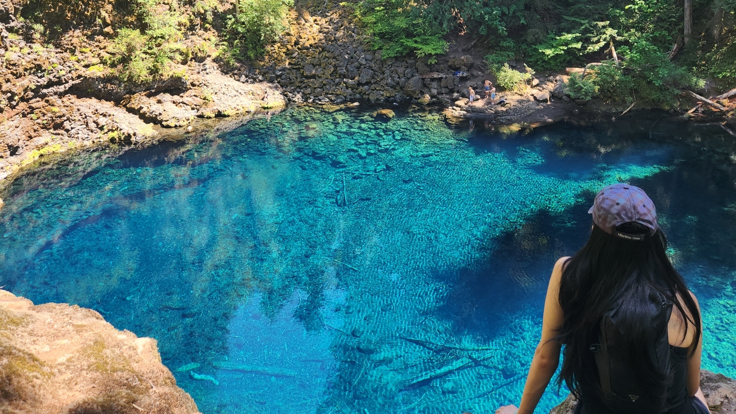 Woman with black hair and hat sits looking at a deep blue pool with trees and rocky shoreline