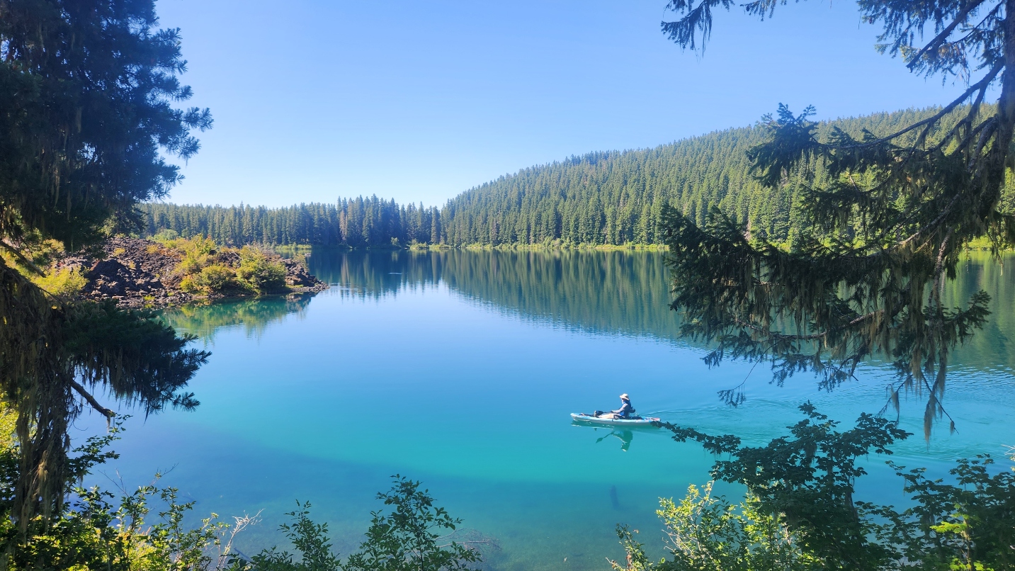 Kayaker paddles on a glassy blue lake with trees and blue sky all around