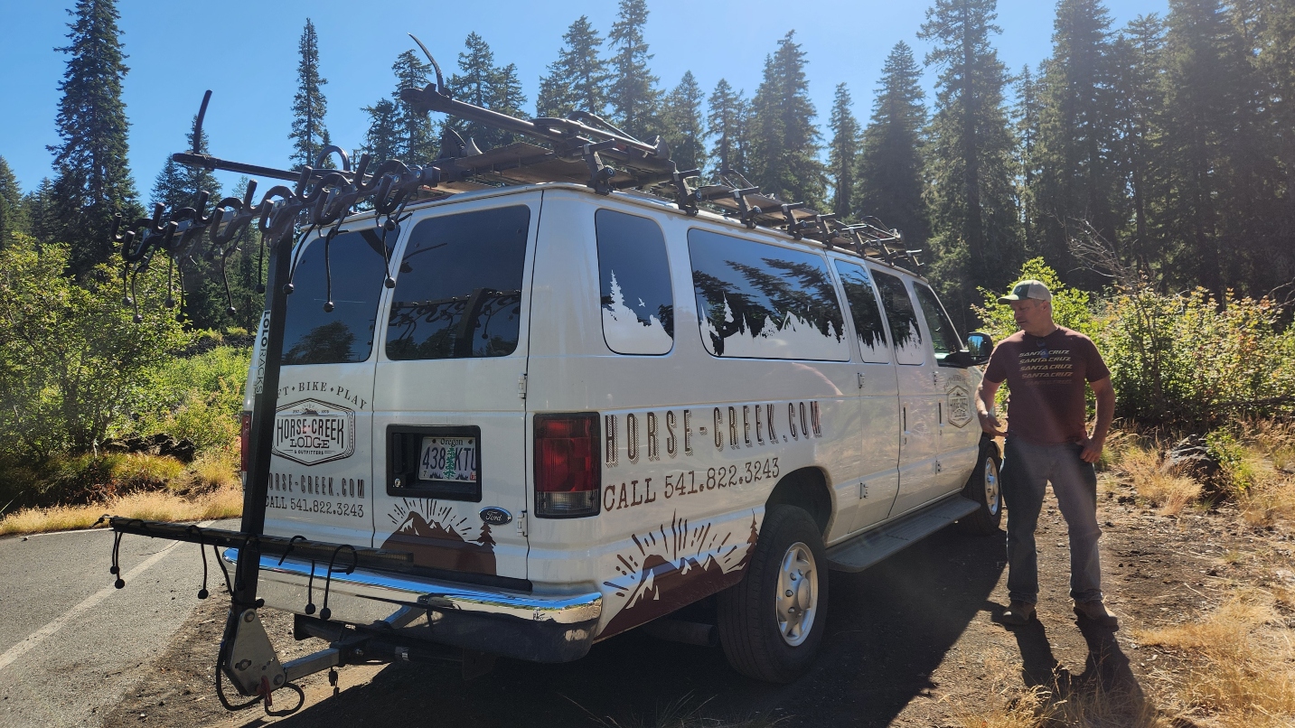 A man wearing T-shirt and jeans stands next to a white van with bike rack and words on the side.