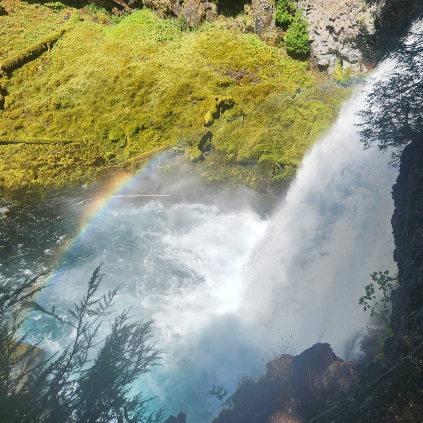 Side view of a waterfall cascading into a pool with a rainbow reflection and mossy rock walls