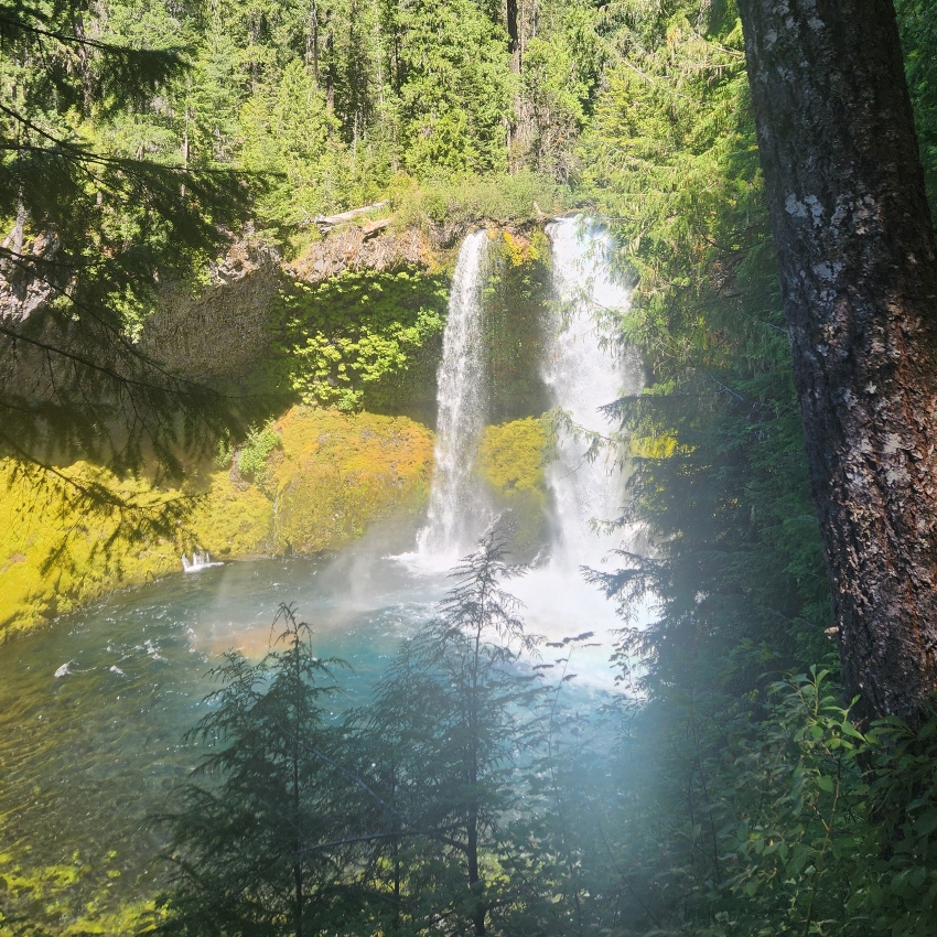 Two cascading waterfalls into a blue pool with a rainbow reflection and trees all around
