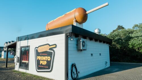 Large hot dog on a stick on top of a small white building with the name "The Original Pronto Pup"
