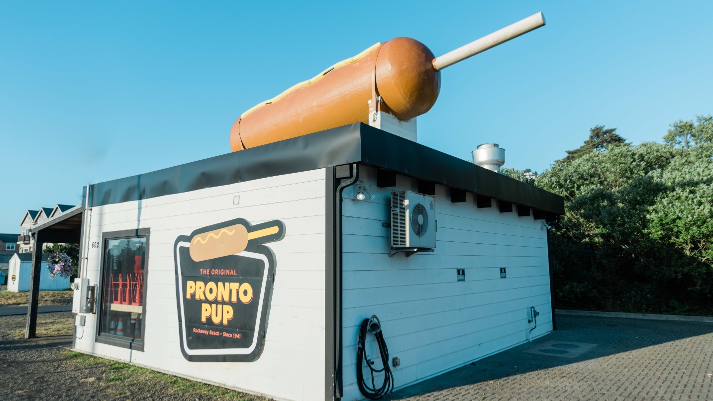 Large hot dog on a stick on top of a small white building with the name "The Original Pronto Pup"