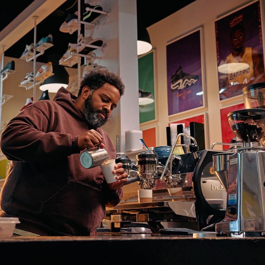 Man with black hair and beard pours coffee from shiny espresso machine, with posters and sneakers in the background