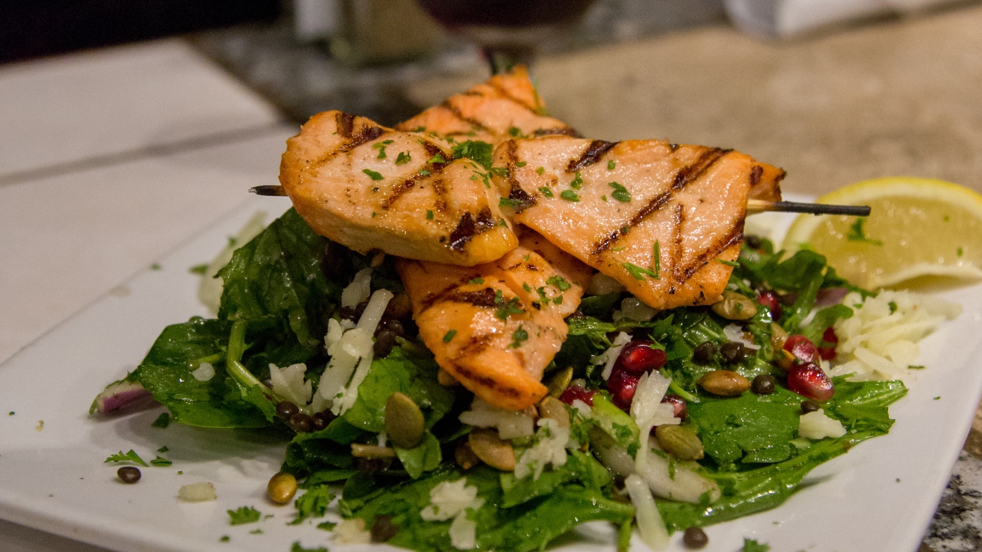 Several pieces of cooked salmon atop a plate of salad with pumpkin seeds, pomegranate seeds and a slice of lemon