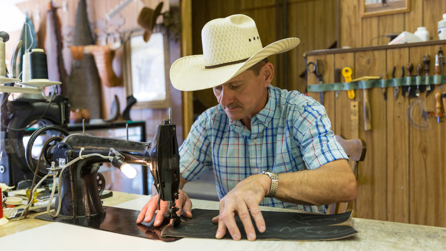 A man in a cowboy hat sewing a piece of leather on an industrial sewing machine.