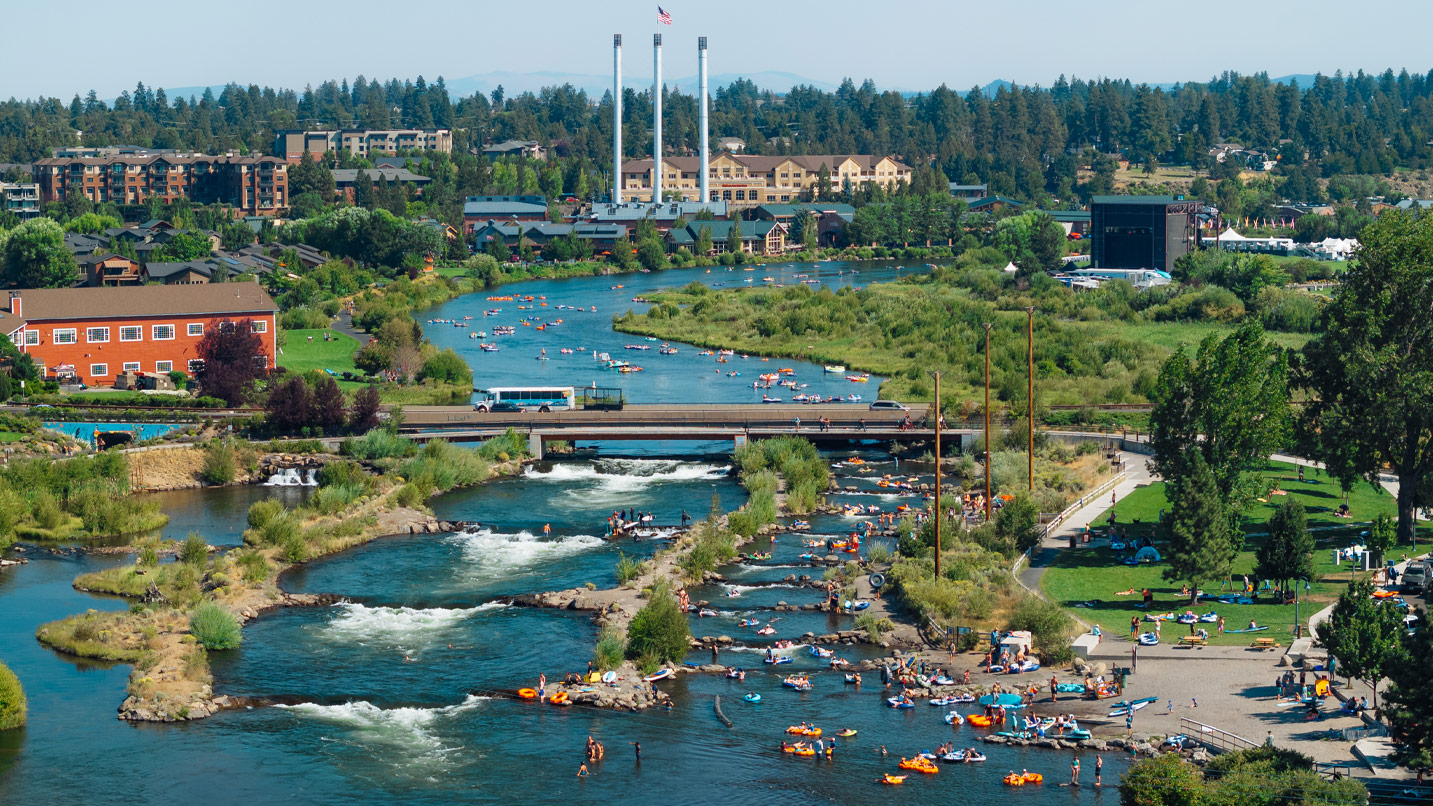 Wide view of the Deschutes River as it cuts through Bend's Old Mill District. Several floaters on inner tubes are seen in the calm part of the river, with views of the historic industrial smokestacks in the distance.