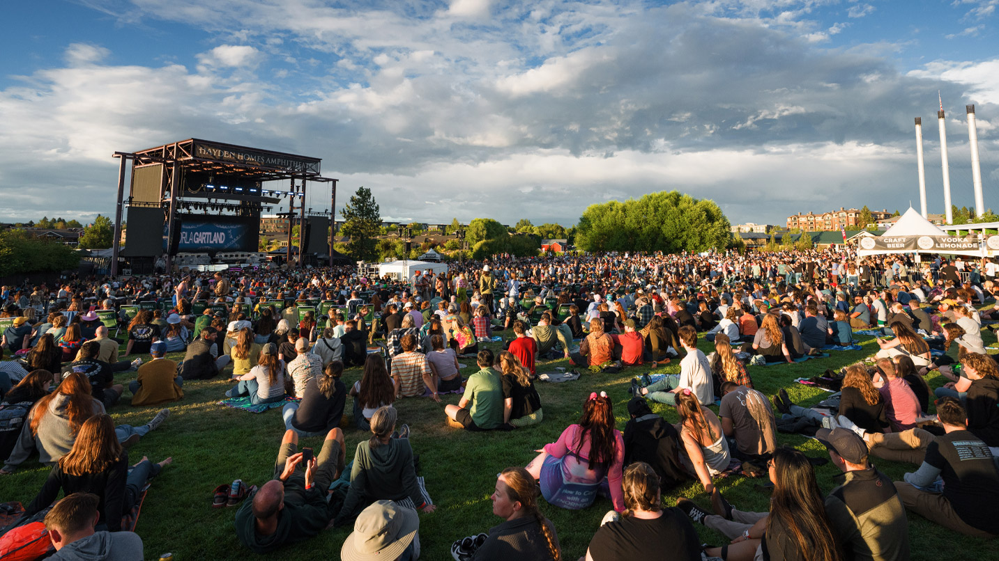 A large crowd of people sitting on a grassy lawn looking towards a large music stage. In the distance, the historic industrial smokestacks from Bend's Old Mill District are visible.