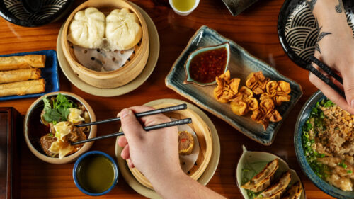 Several plates of Asian cuisine, including lumpia, baos in a steam basket, fried rangoons and a noodle dish. Two hands with chopsticks reach for food on the plates.