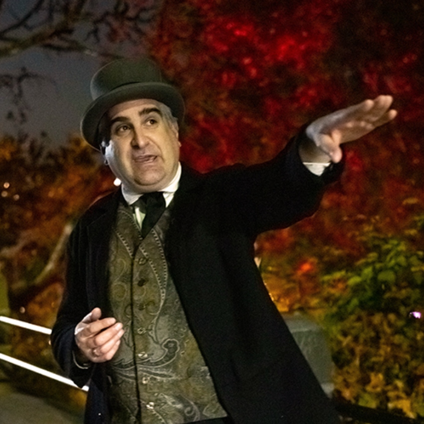 Man wearing top hat, vest, tie and dark coat pointing to something against a night sky