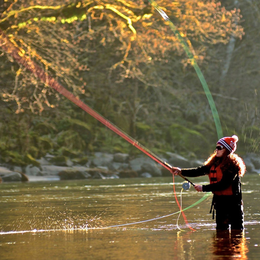 Woman standing in river wears sunglasses, red beanie and red PFD and casts a fishing line
