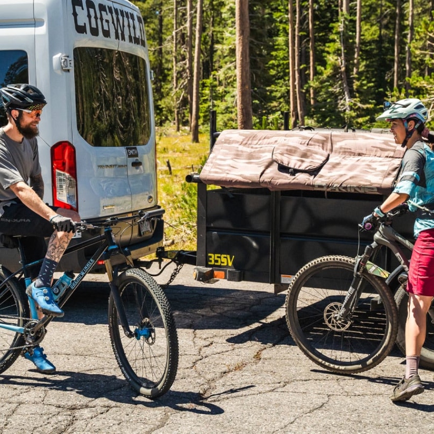 Man with bicycle and woman with bicycle smile, with white Cog Wild van and trailer in background 