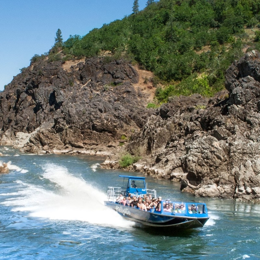 Blue boat full of people jets down a river with large rocky bluff and blue sky in background