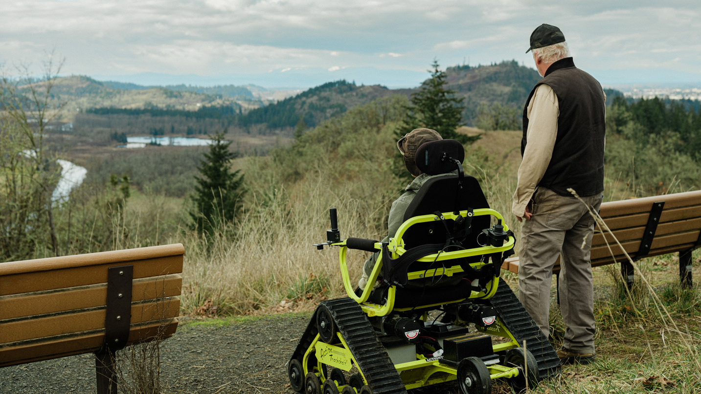 Two men, one standing and the other in an all-terrain wheelchair, overlooking a view from Pisgah Arboretum.