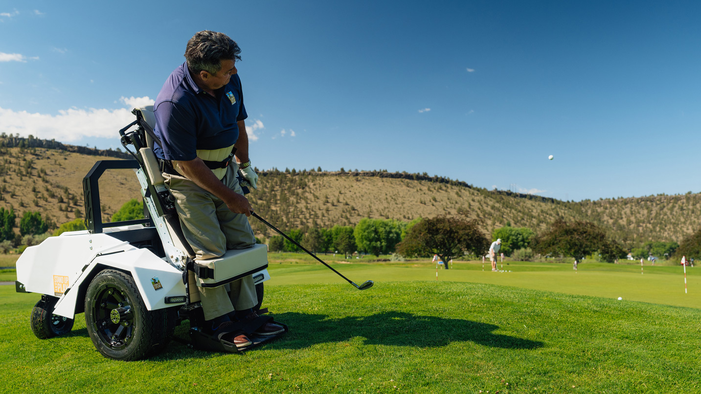 A man in an adaptive golf cart, which helps him stand upright as he tees off on a golf course.