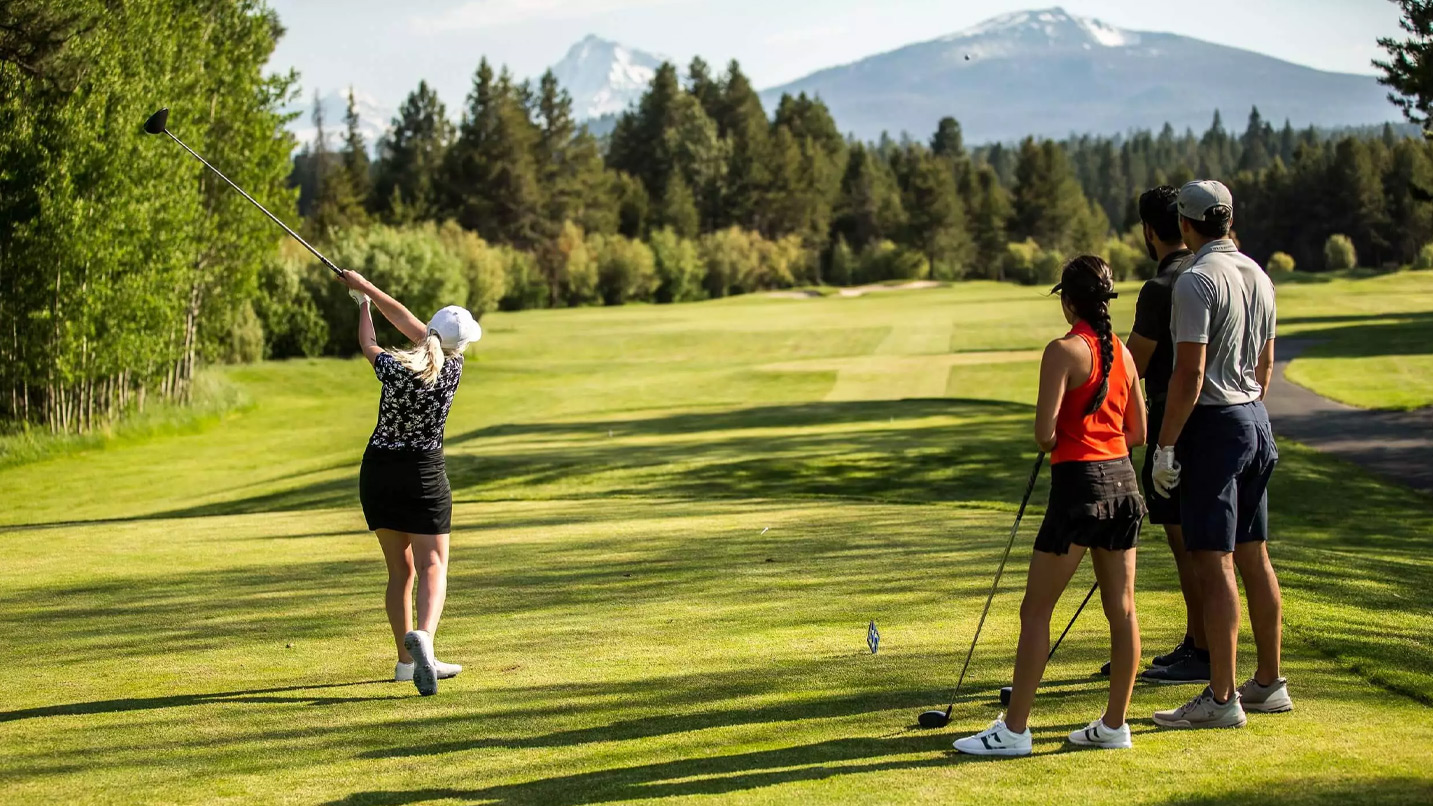 A woman golfer in mid-swing on a golf course with a mountain view in the background. Another woman and two men stand to the side watching.
