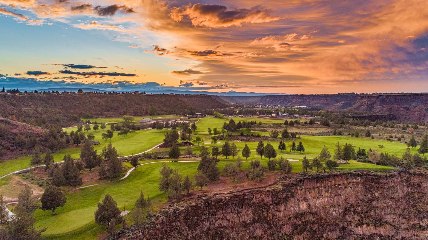 An aerial view of a golf course and it's scenic surroundings, which include cliffside views and orange and purple clouds during sunset.