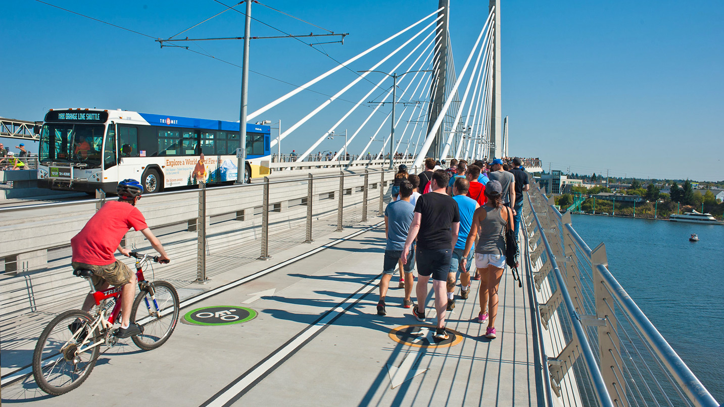 A large group of people walk on a pedestrian path of the Tilikum Bridge. A single cyclist rides on the bike lane, while a TriMet bus drives through the center of the bridge.