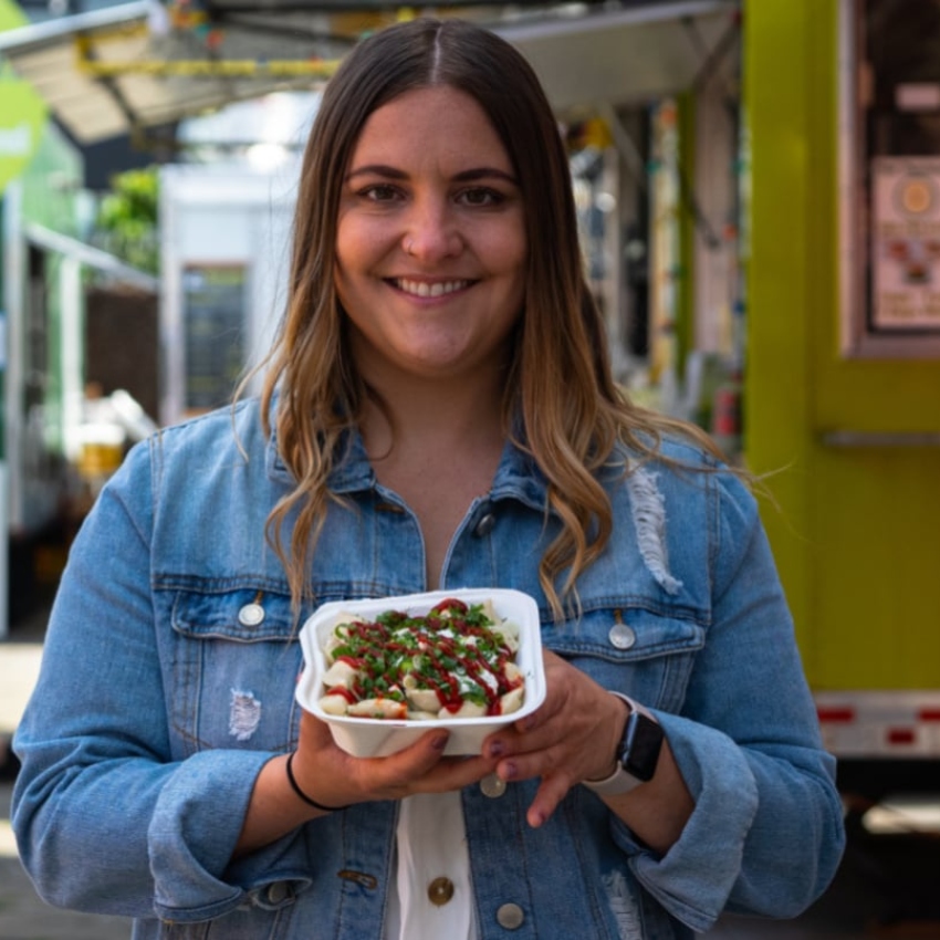 Woman wearing blue button-down shirt smiles as she holds takeout dish of food toward camera in front of food cart