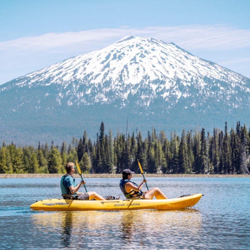 Snowcapped mountain in background and two people paddling a yellow kayak on a lake in the foreground 